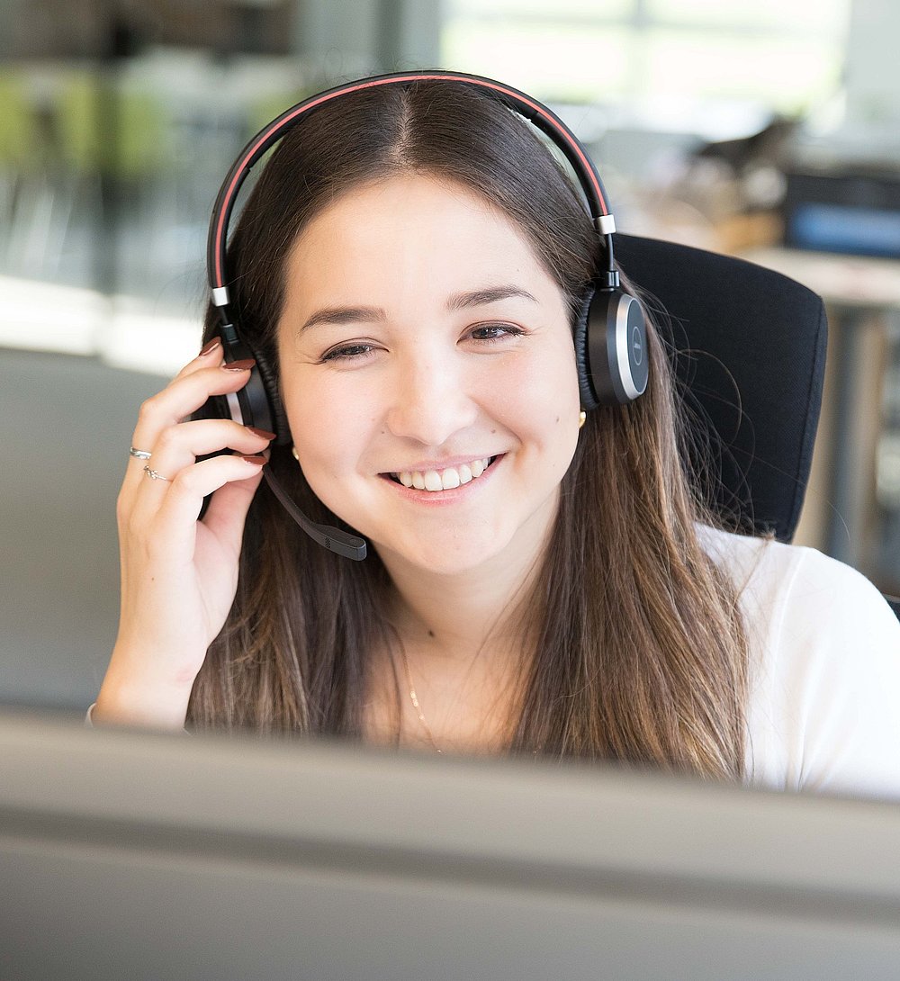Eine freundliche junge Frau mit Headset arbeitet lächelnd an einem Computer in einem modernen Büro. Sie wirkt konzentriert und kommunikativ, als führe sie gerade ein Gespräch oder unterstütze Kund*innen.